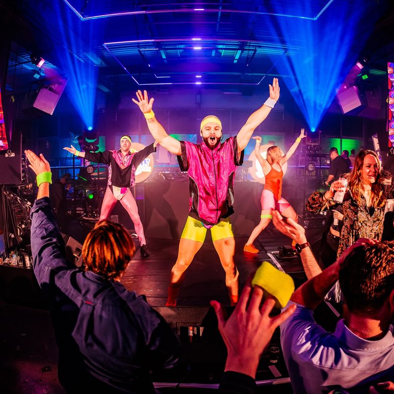 Three performers in neon aerobics outfits on stage, the crowd with their hands in the air